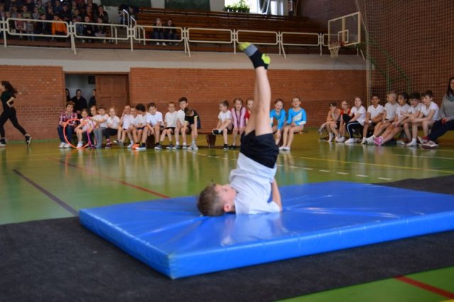 Talajtorna verseny alsósok A képen egy tornateremben egy gyerek talajtorna gyakorlatokat végez egy szőnyegen, a háttérben és a lelátón gyerekek láthatóak.