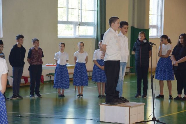 Témahét - Kossuth hét A képen egy dobogón áll és szaval két gyerek, körben gyerekek állnak. 